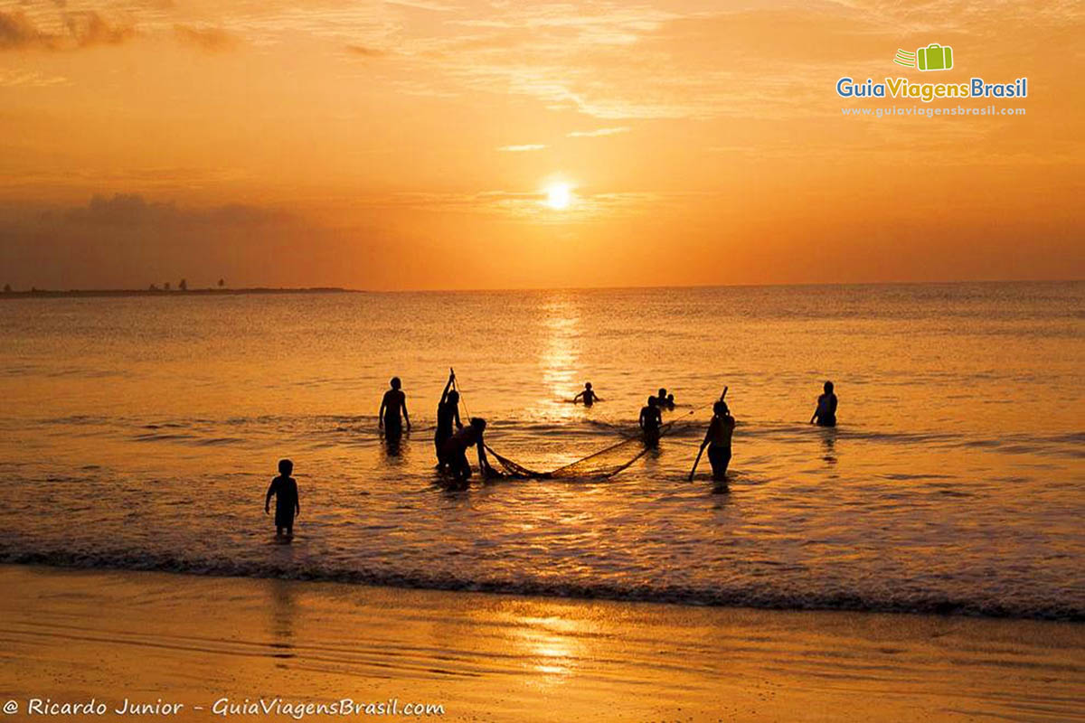 Imagem de pescadores puxando a rede na beira praia e lindo céu alaranjado.