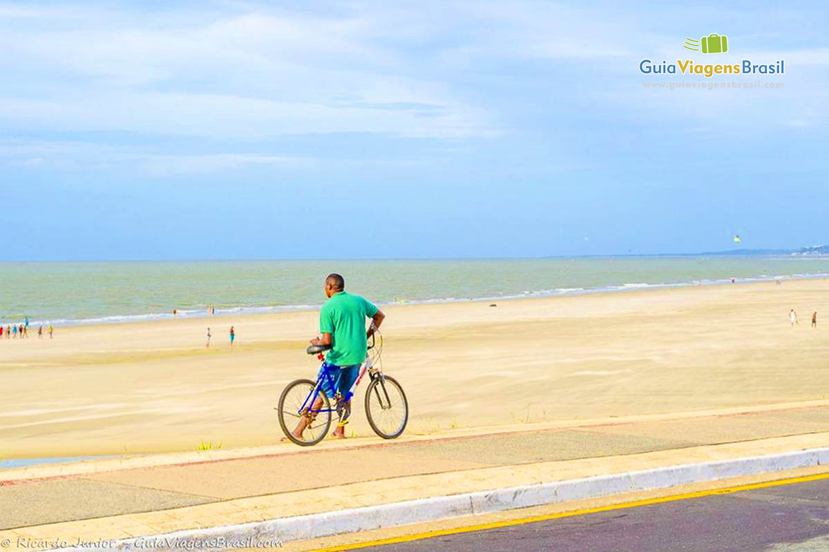 Imagem de um rapaz parado com sua bicicleta no calçadão, admirando a praia.