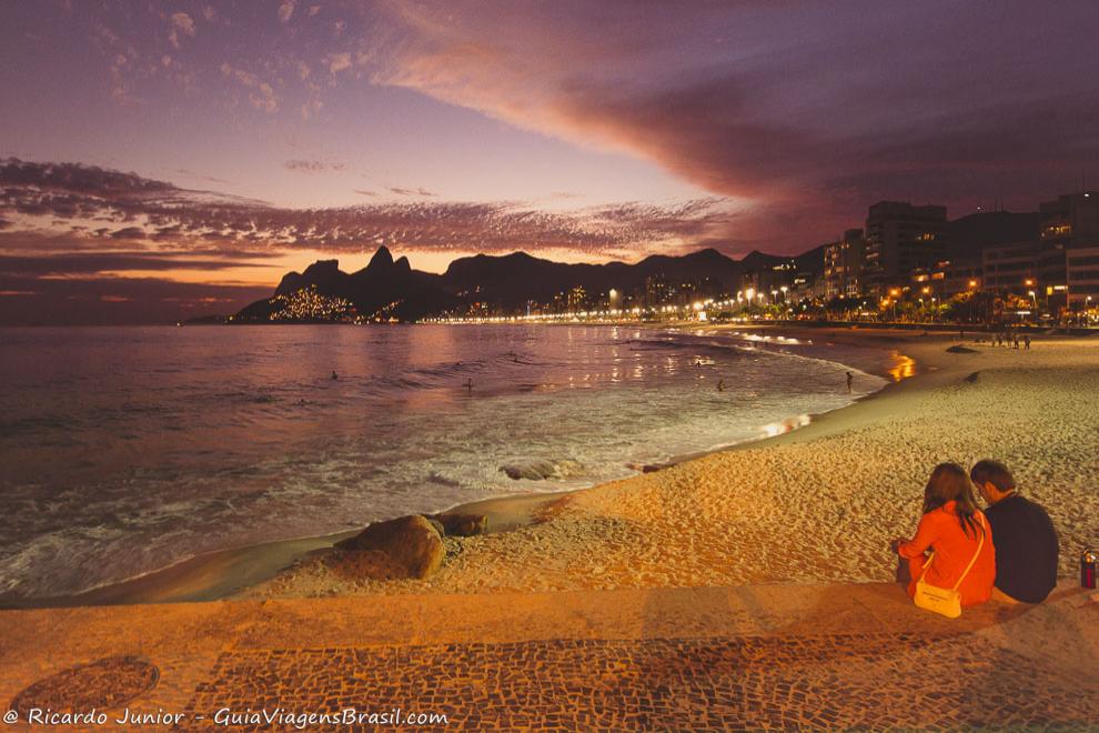 Imagem de casal admirando o belo entardecer com a praia já iluminada no Arpoador.