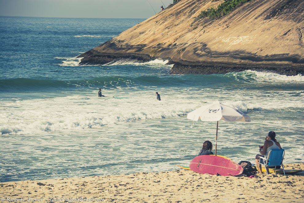 Imagem de amigos sentados na areia vendo surfistas e admirando a Praia do Recreio dos Bandeirantes.