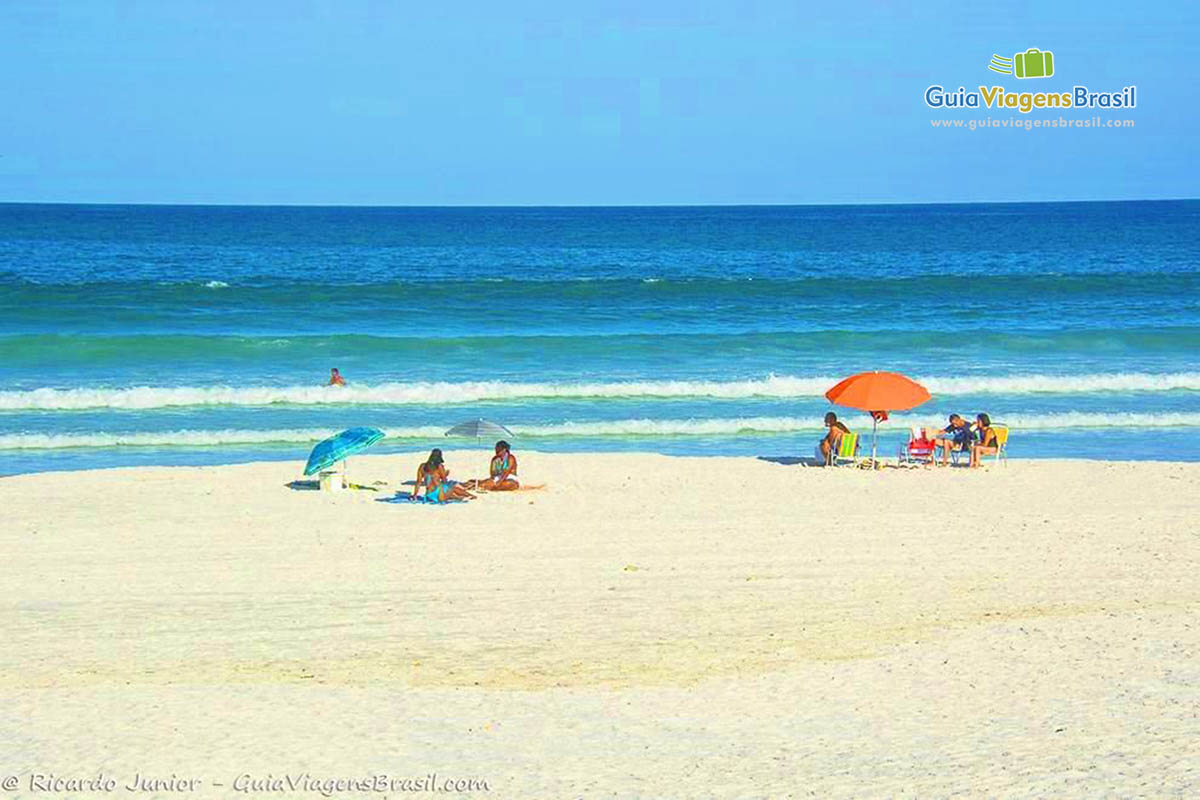 Imagem de amigos e amigas sentados na areia admirando a Praia das Dunas.