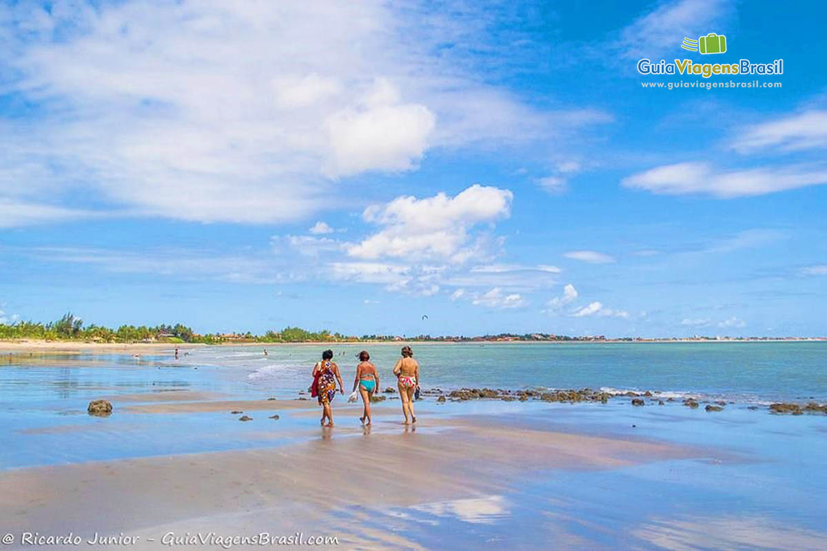 Imagem de três amigas caminhando na beira da praia.