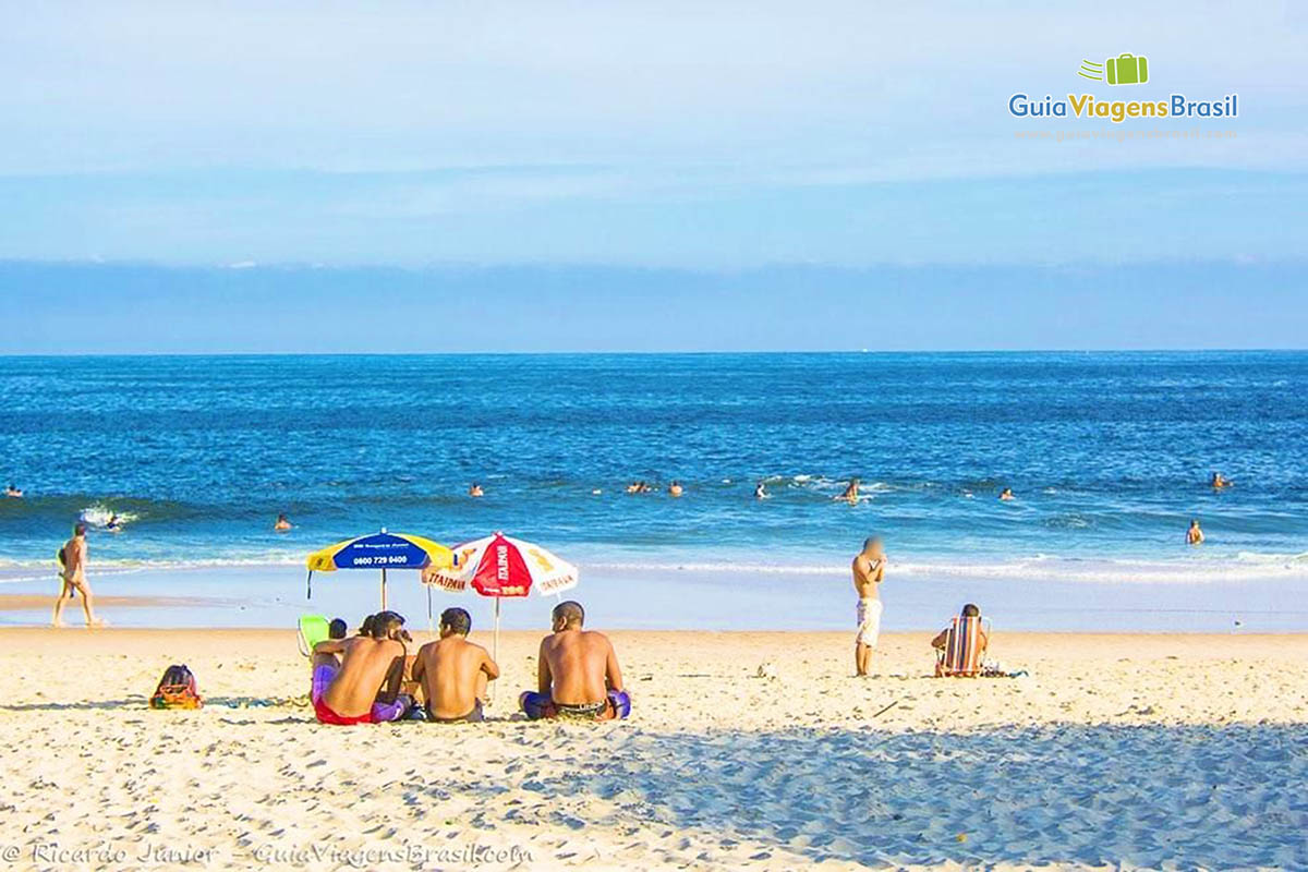 Imagem de três amigos sentados embaixo de guarda sol olhando em direção ao mar.