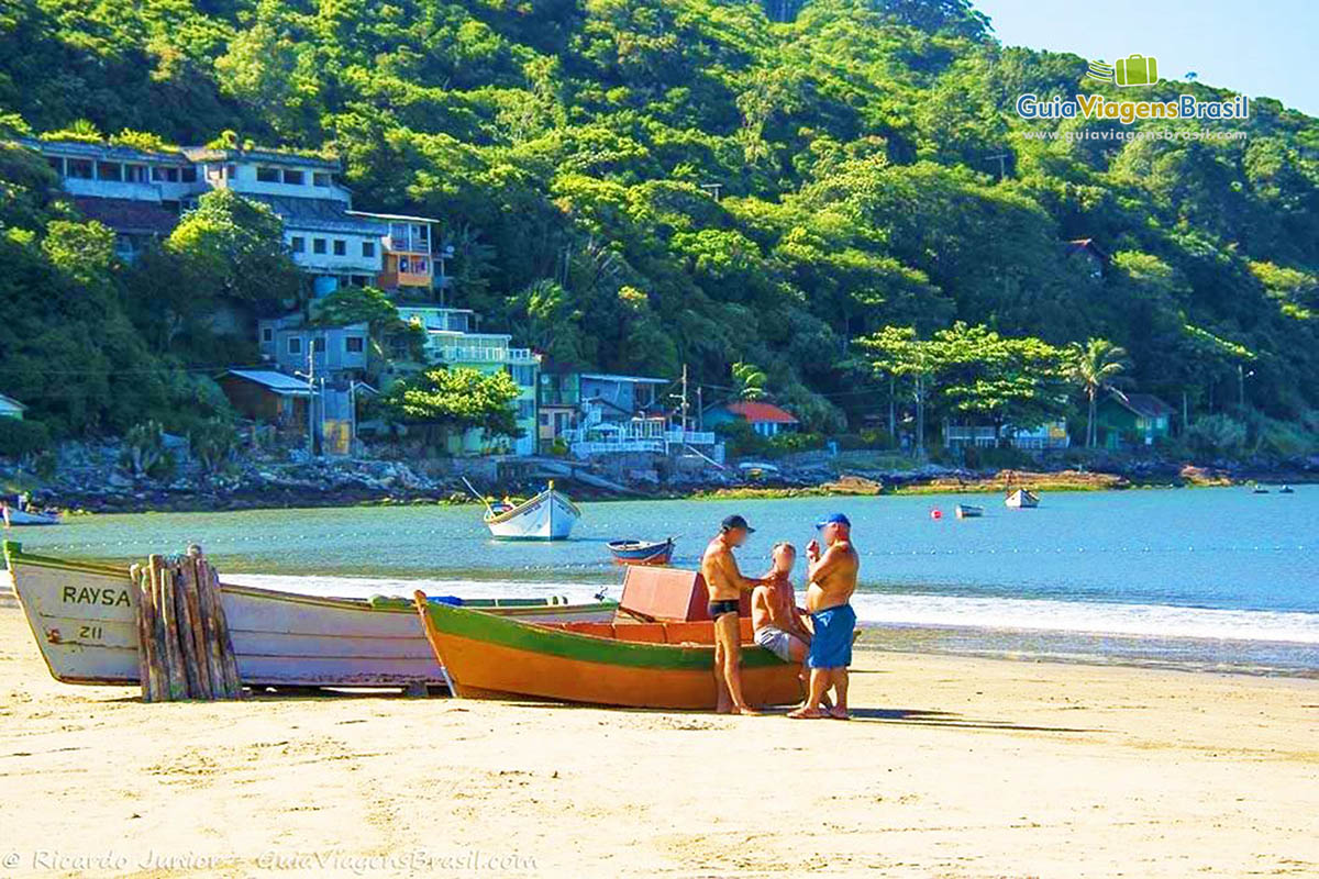 Imagem de três senhores conversando encostados em um barco de pescador que está na areia da praia.