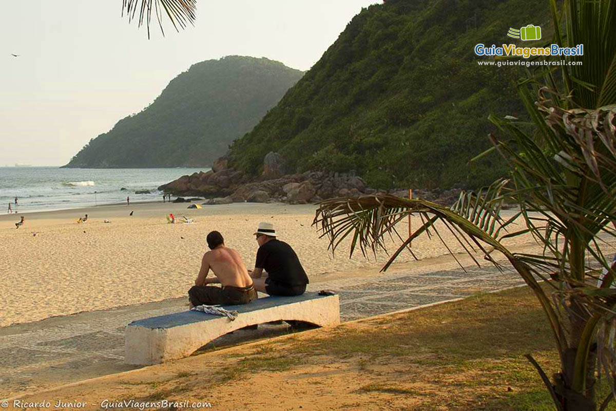 Imagem de amigos sentados no banco de cimento nas areias da Praia do Tombo no Guarujá.