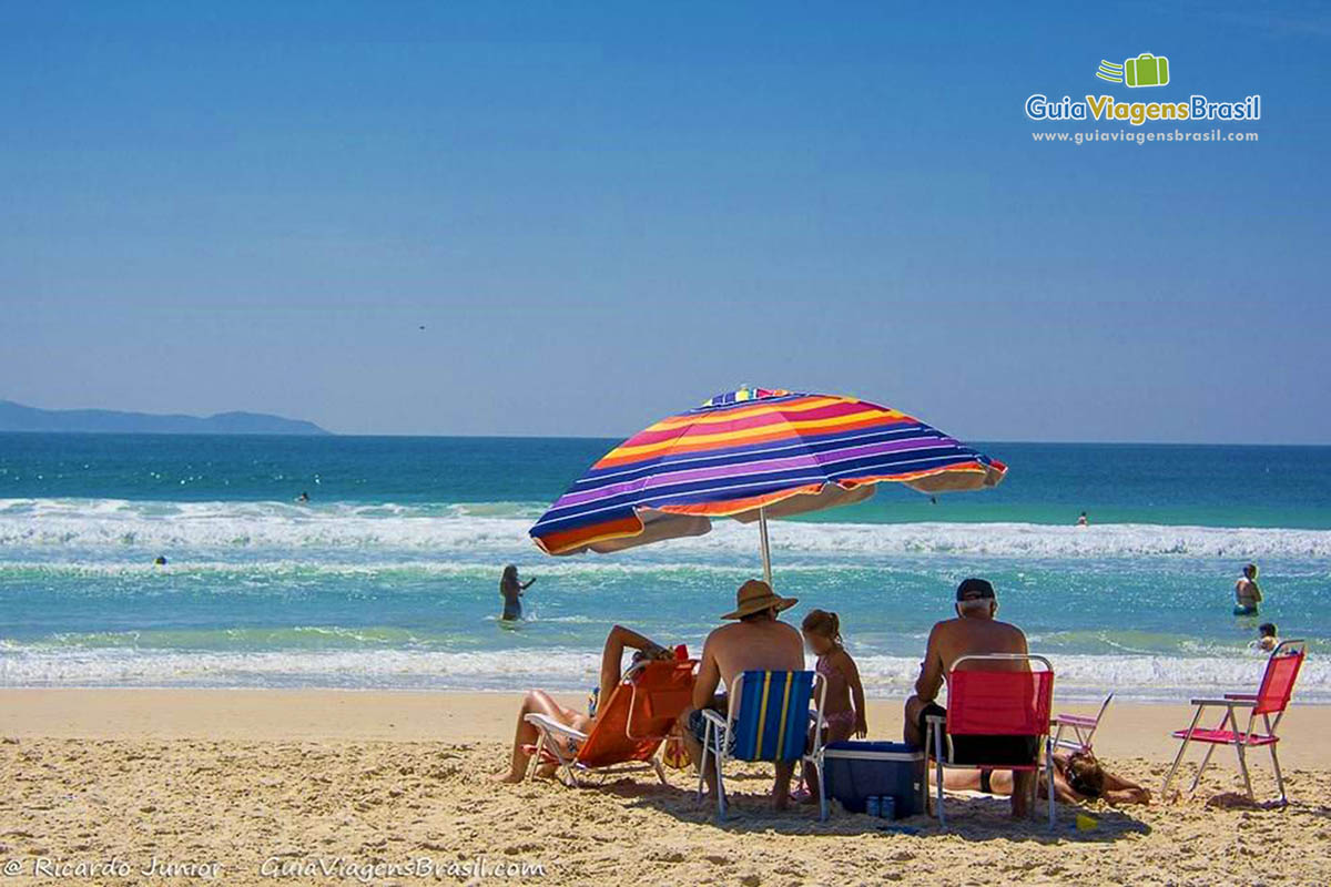 Imagem de uma família sentados em baixo do guarda sol e aproveitando um belo dia de sol.