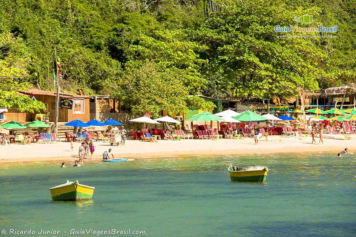 Imagem do barcos no mar e ao fundo a areia e os quiosques que possui na praia. 