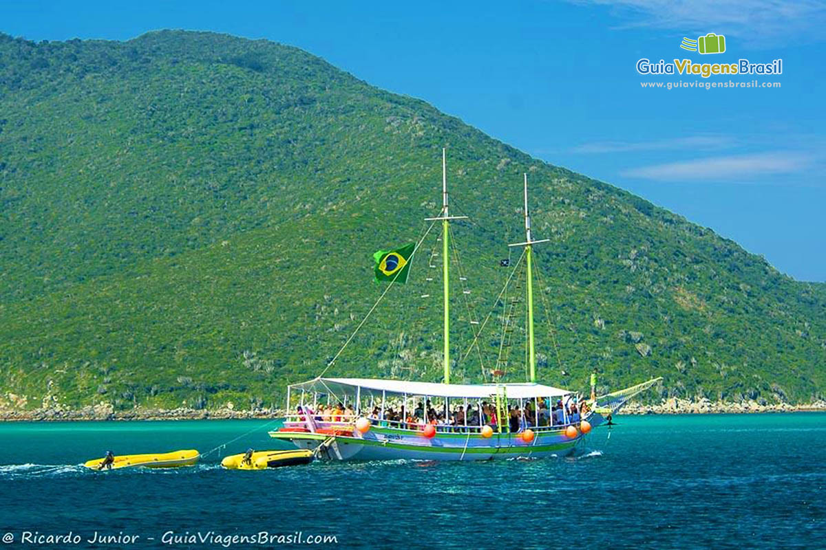 Imagem de barco de passeio repleto de turistas com a bandeira do Brasil e puxando dois botes.