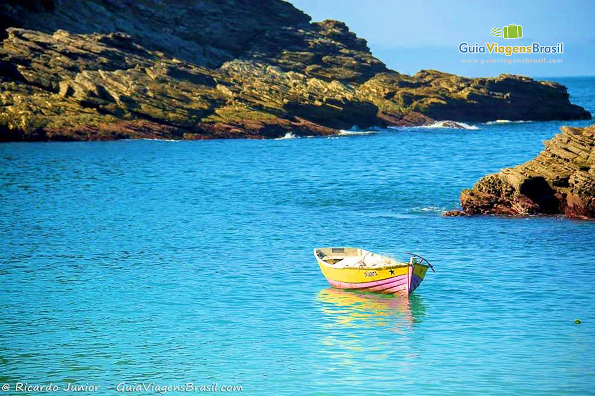 Imagem de barco de pescador no mar azul e ao fundo pedras da praia.
