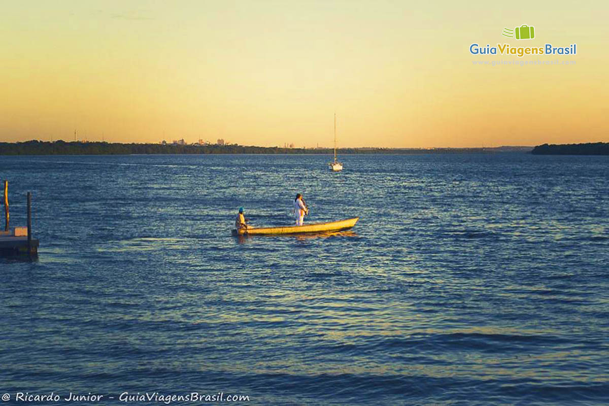 Imagem de uma moça em pé em uma canoa e um senhor remando, no entardecer da Praia Jacaré, em João Pessoa.