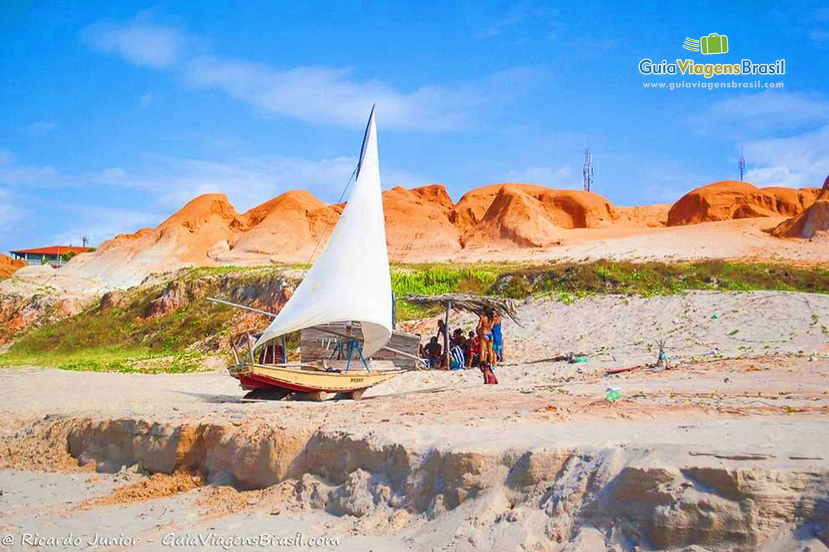 Imagem de barquinho a vela na areia da praia ao lado de barraca de sapê.