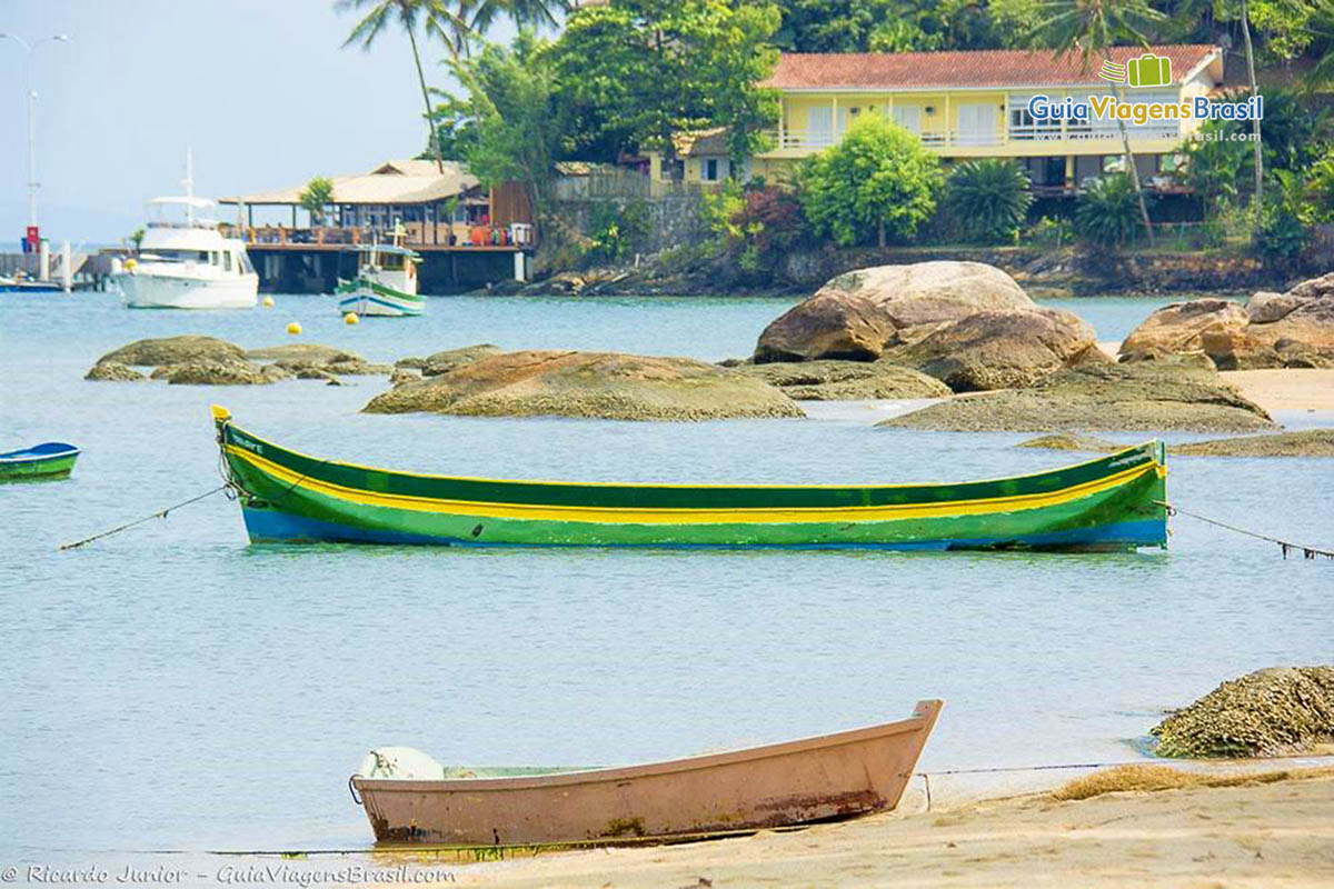 Imagem de uma canoa verde e amarela nas águas e ao fundo umas pedras e linda casa no morro.