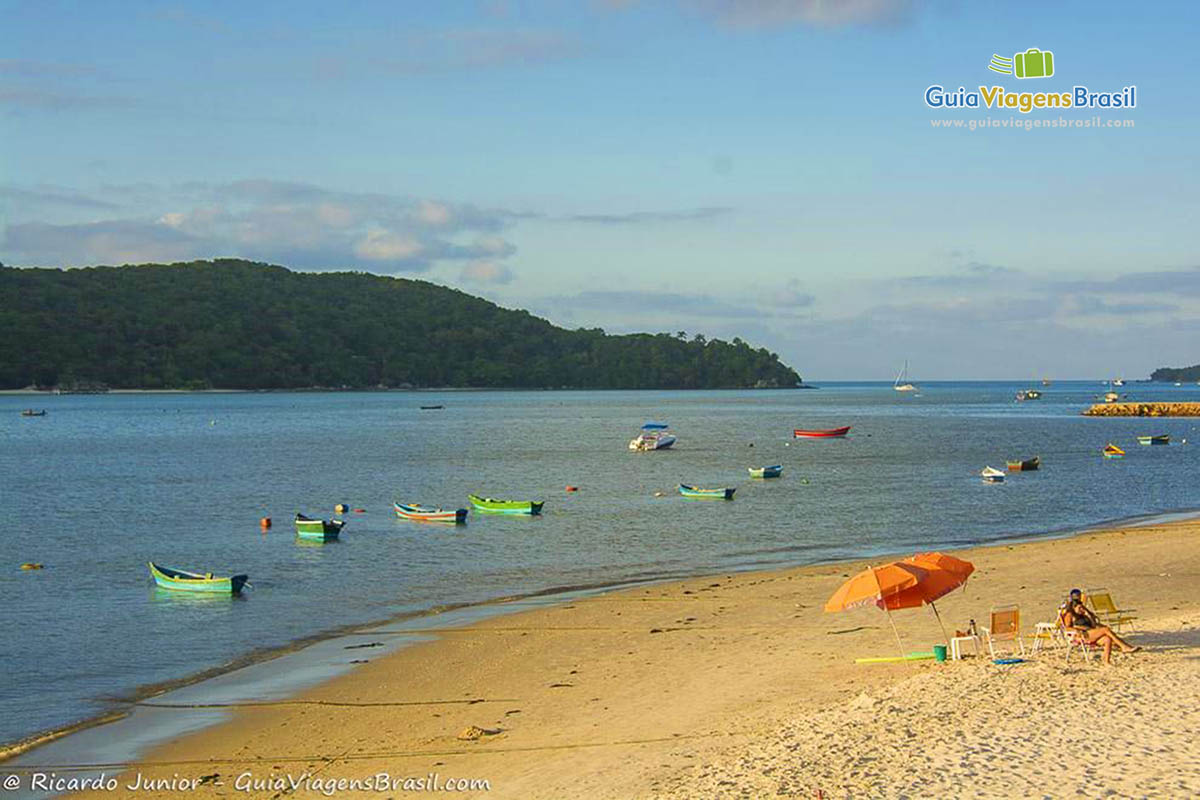 Imagem de um guarda sol próximo ao mar e barcos pequenos de pesca na beira do mar.