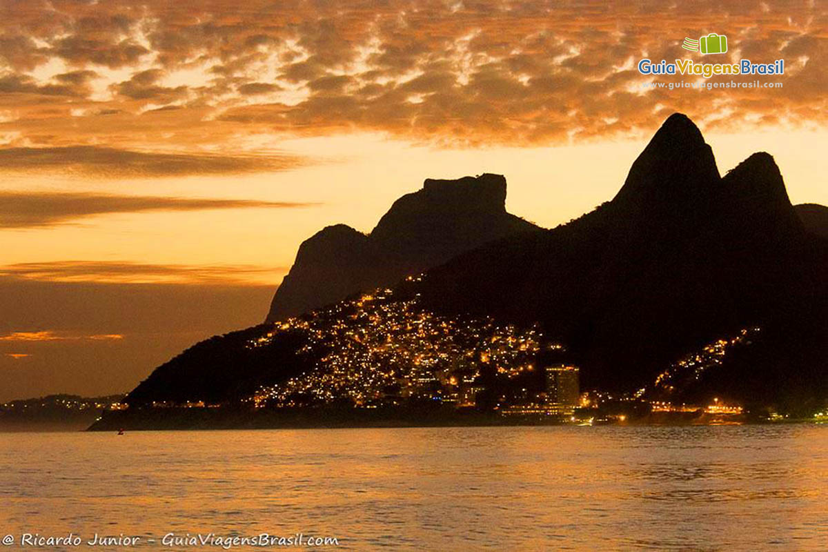 Imagem das águas do mar e ao fundo morro já com as casas todas iluminadas, ressaltando a beleza do local.