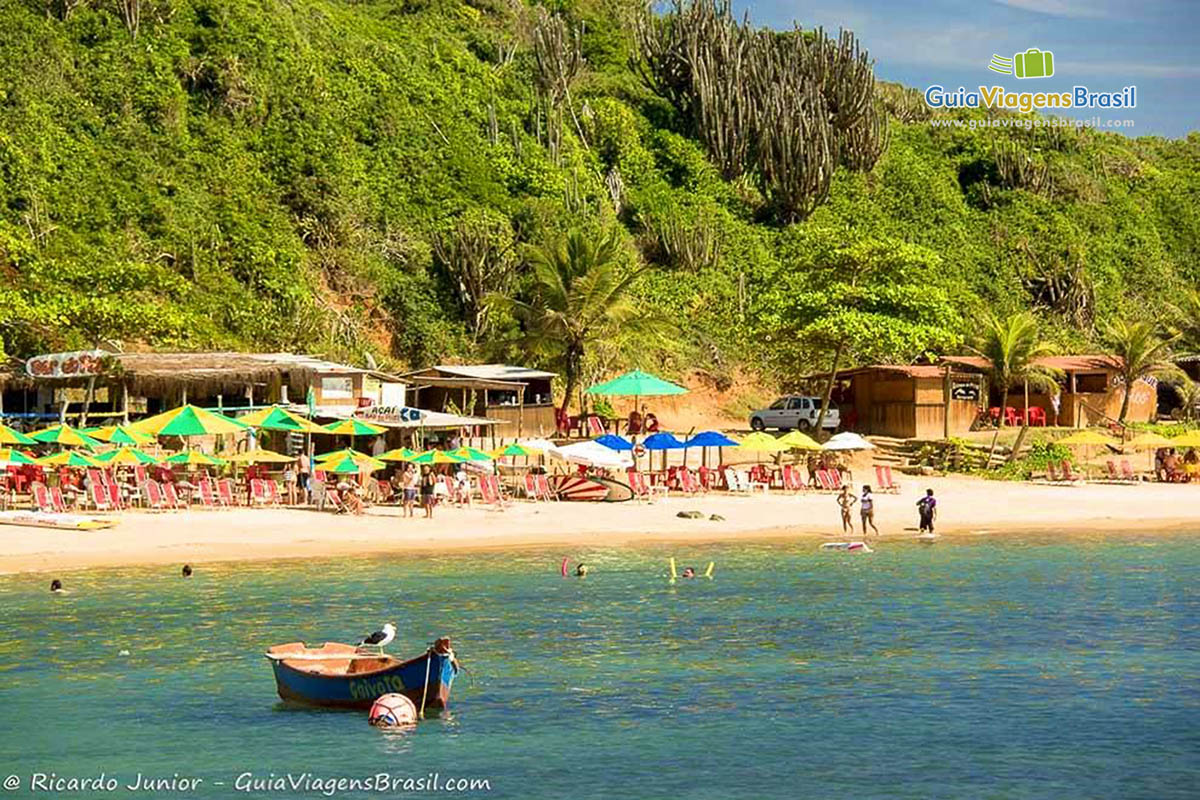 Imagem do mar e ao fundo nas areias cadeiras e guarda sol já arrumados para receber visitantes da Praia Tartaruga.