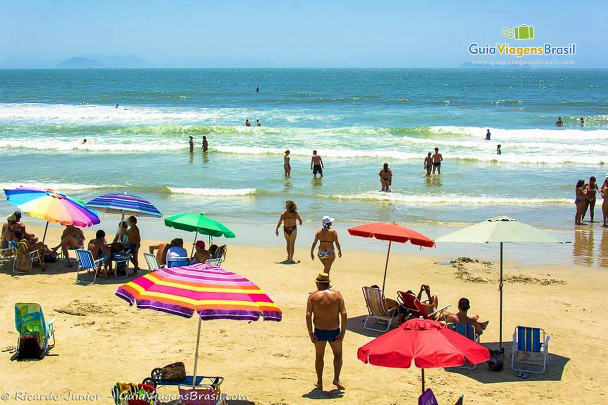 Imagem de guarda sol próximo a beira da praia e pessoas no mar.