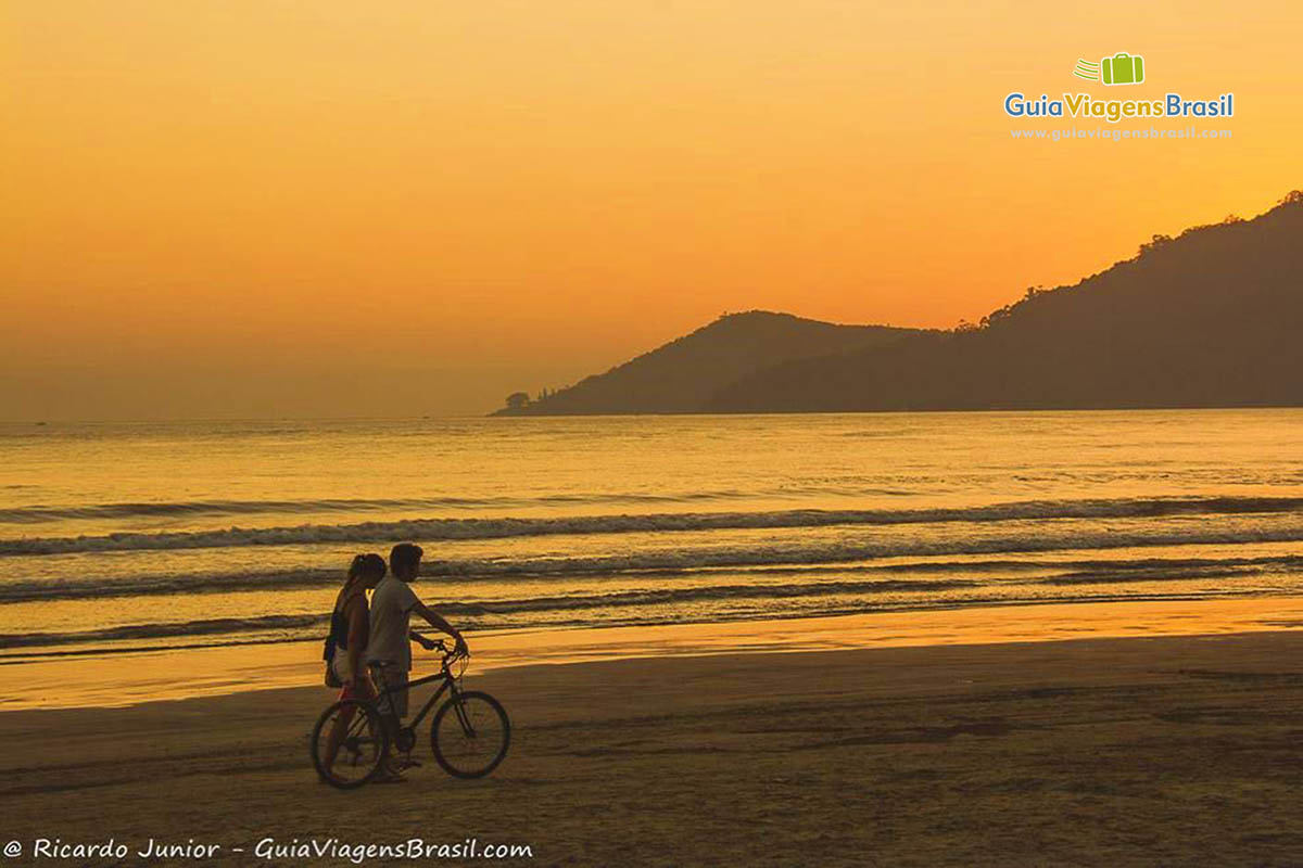 Imagem de duas pessoas empurrando bicicleta na beira do mar e ao fundo o céu alaranjado de um lindo entardecer.