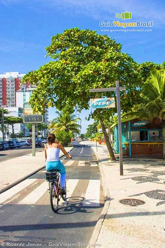 Imagem de uma mulher andando de bicicleta na ciclovia, na Praia Barra da Tijuca.