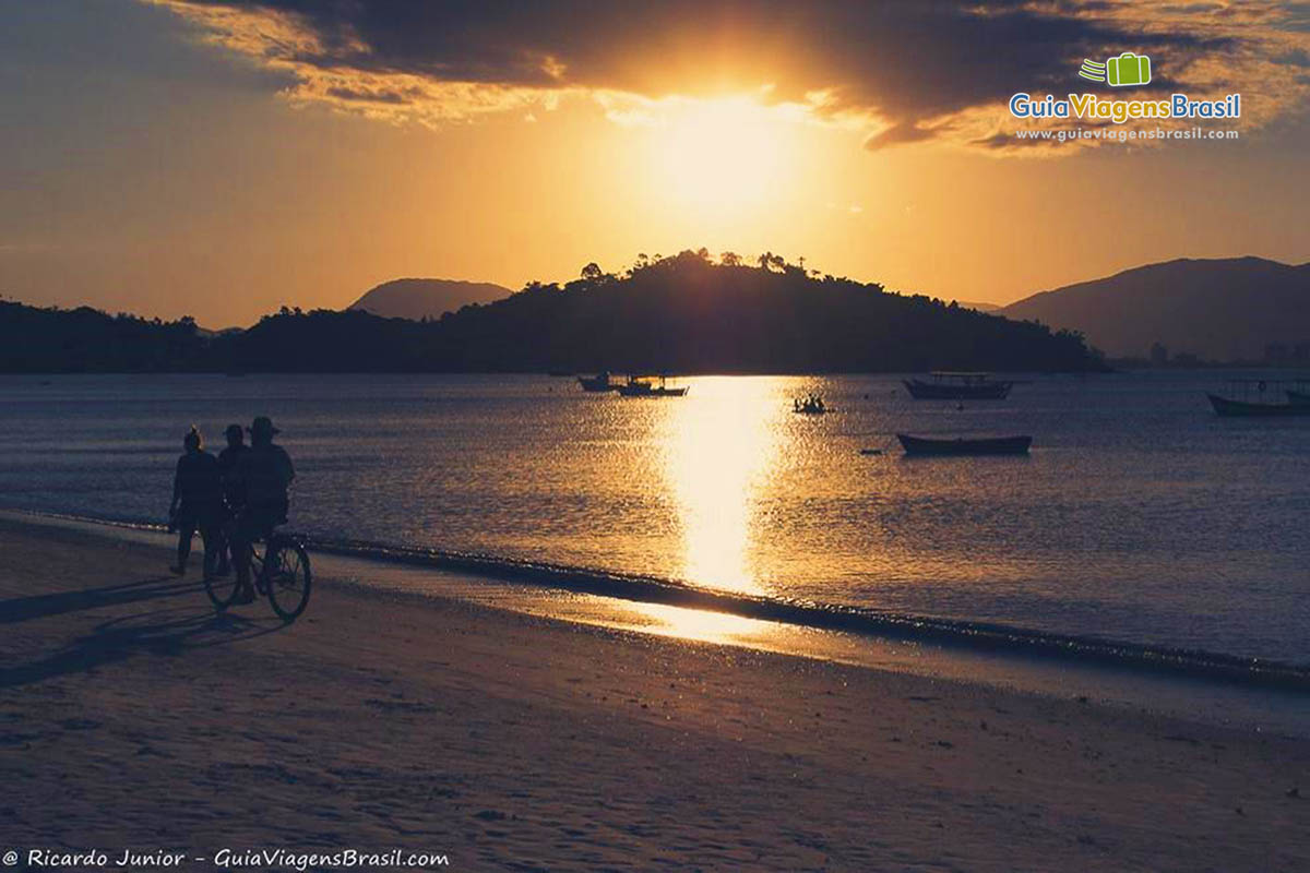 Imagem de pessoas caminhando e andando de bicicleta na praia e ao fundo belo por do sol.