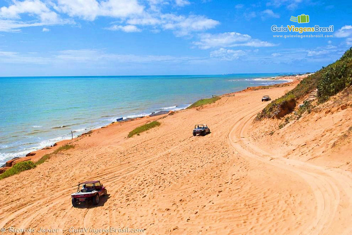 Imagem de vários bugues andando pelas falesias avermelhadas e ao fundo o mar da Praia Canoa Quebrada.