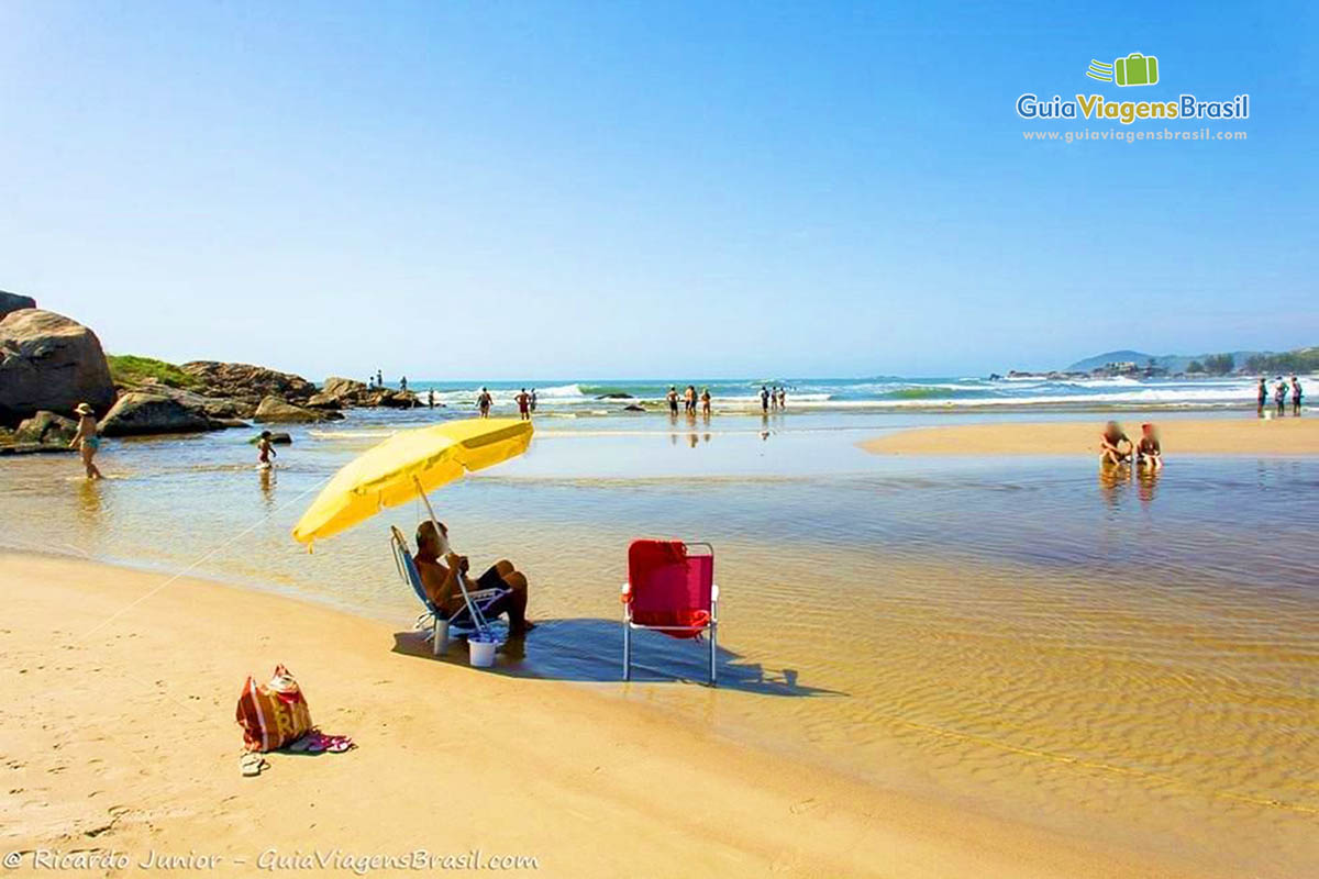 Imagem de uma pessoa sentada em baixo do guarda sol e algumas pessoas no canto da praia.