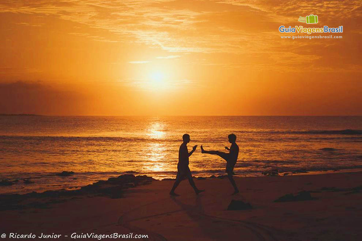 Imagem de duas pessoas jogando  capoeira no belo fim de tarde na praia.
