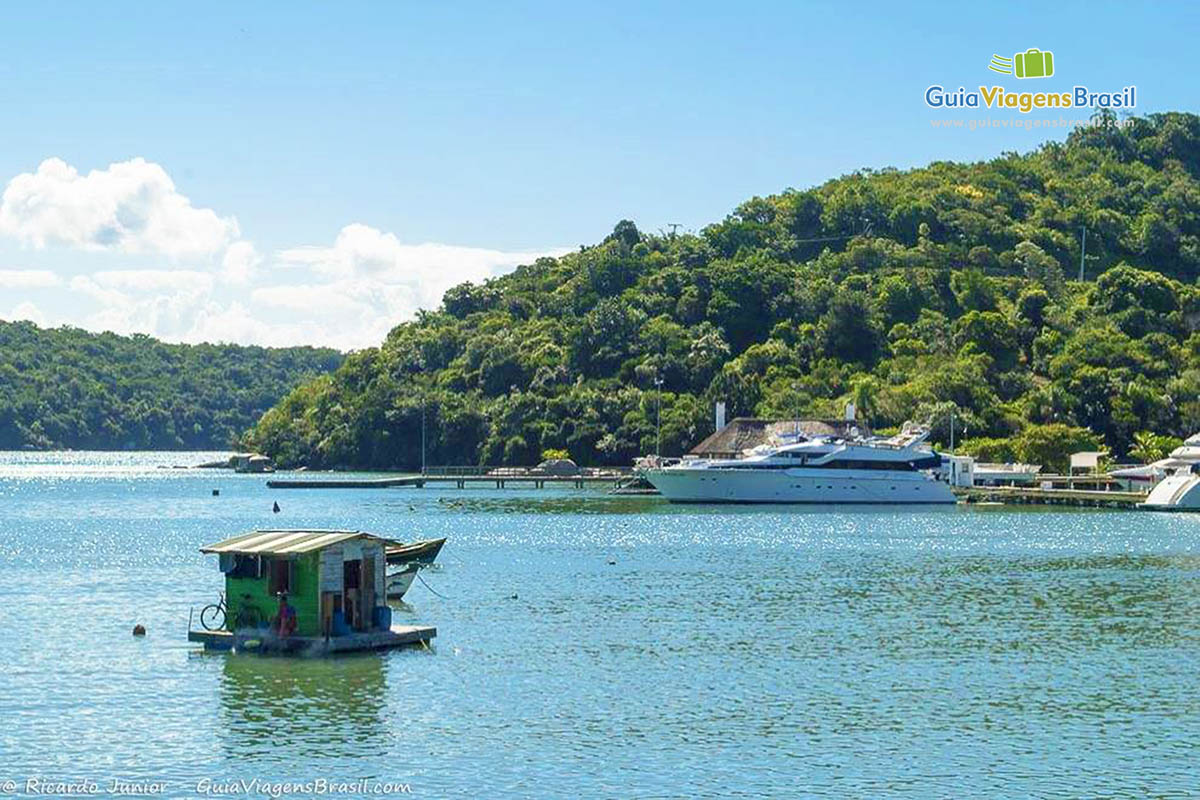 Imagem de uma pequena casa flutuante e ao fundo uma luxuosa lancha na Praia Central.