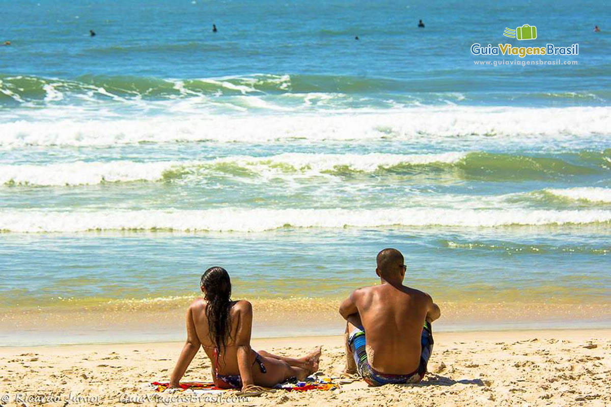 Imagem de casal sentados na areia admirando o belo mar.