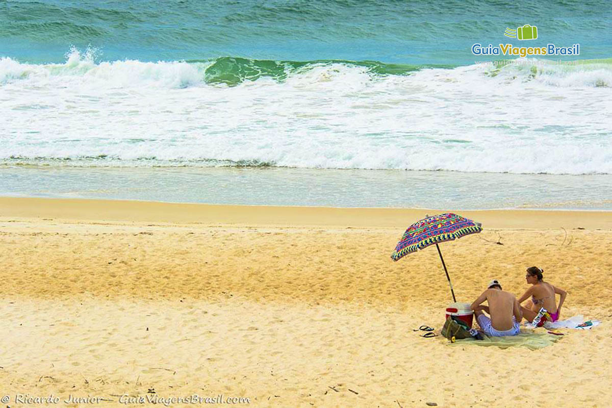 Imagem de casal sentados na cadeira embaixo guarda sol na bela praia.
