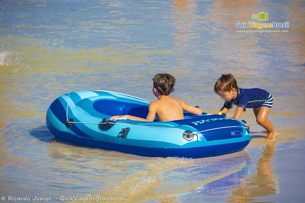 Imagem de um menino empurrando uma outra criança dentro da bóia, na beirinha do mar.