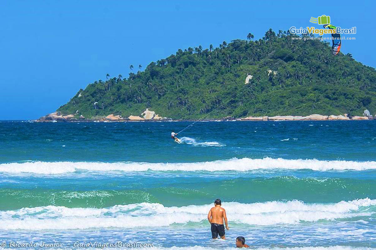 Imagem de crianças na beira da praia e uma pessoa praticando kitesurf ao fundo.