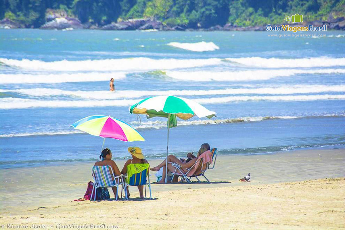 Imagem de pessoas sentados embaixo de seu guarda sol conversando e curtindo a praia.