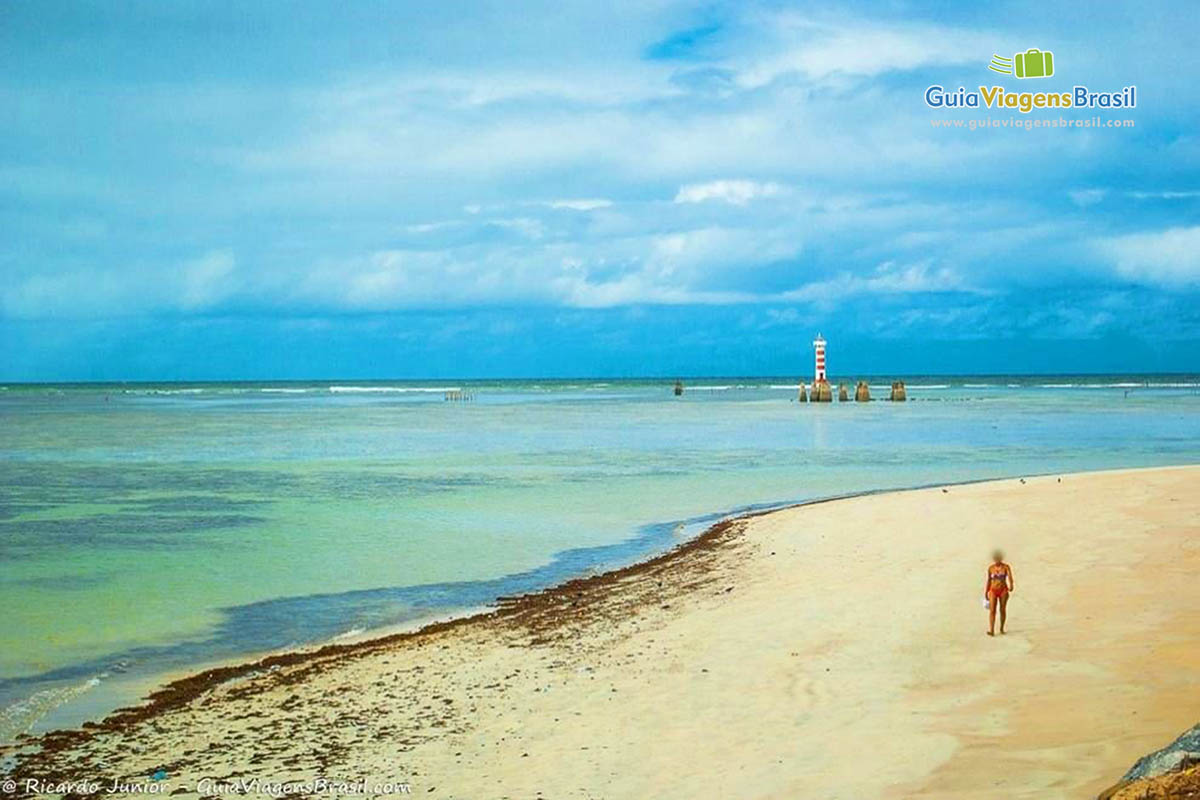 Imagem de pessoa andando nas areias e ao fundo lindo farol.