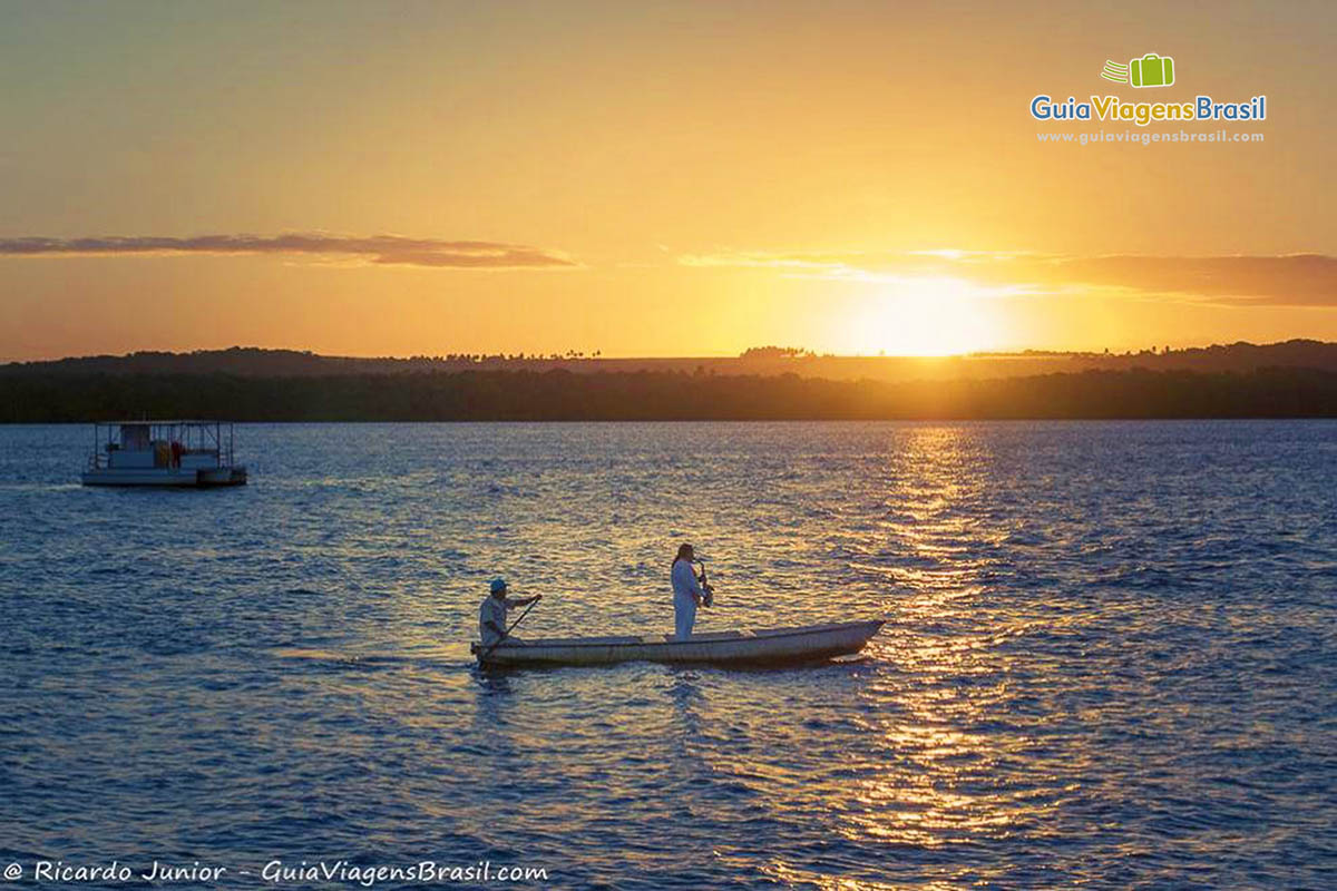 Imagem de dois barcos no mar e ao fundo o sol se pondo na cor alaranjada.