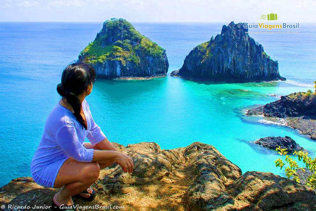 Imagem da turista nas pedras admirando do alto a Baía dos Porcos em Fernando de Noronha, Pernambuco, Brasil.