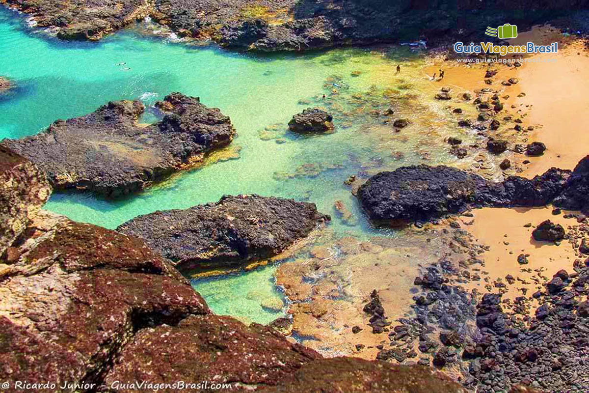 Imagem aérea da Praia Baía dos Porcos, sendo possível ver dois turistas mergulhando entre as pedras e poucas pessoas na areia, em Fernando de Noronha, Pernambuco, Brasil.