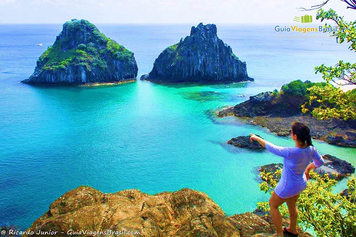 Imagem de uma turistas em pé, apontando para um barco bem distante saindo de trás das pedras, na Baía dos Porcos, em Fernando de Noronha, Pernambuco, Brasil.