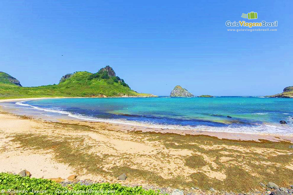 Imagem da praia e ao fundo pequeno morro com vegetação bem verde, na Baía Sueste, em Fernando de Noronha, Pernambuco, Brasil.