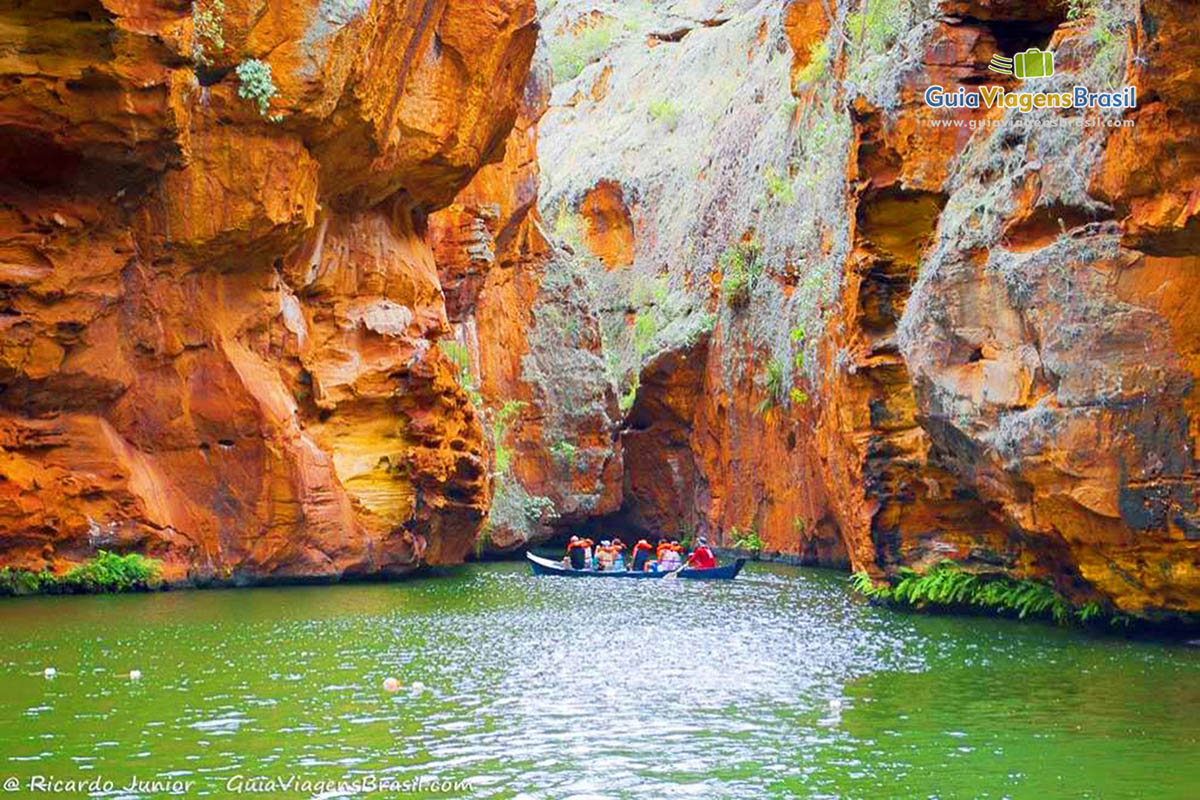 Imagem aproximada das rochas avermelhadas e turistas no barco.