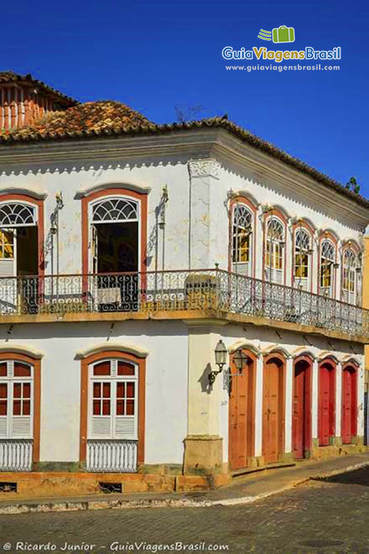 Imagem de casarão de esquina com portas vermelhas e janelas brancas, sendo que as janelas superior estão abertas.