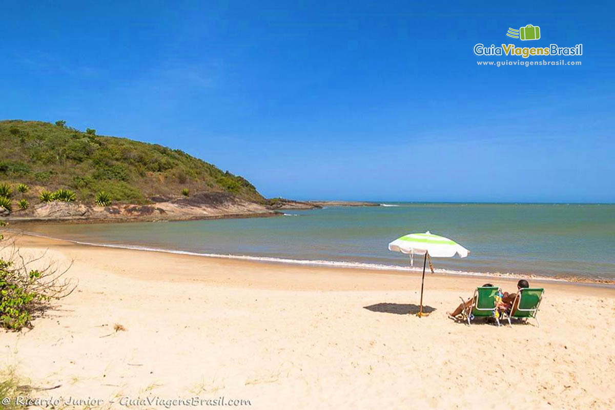 Imagem de duas pessoas sentadas em cadeiras verdes e com guarda sol, curtindo a tarde na praia.