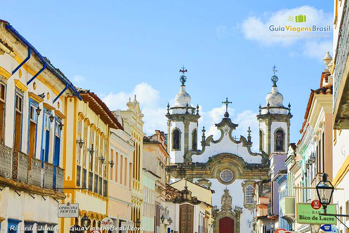 Imagem da arquitetura das casas e o comércio da rua central e ao fundo a fachada da igreja.