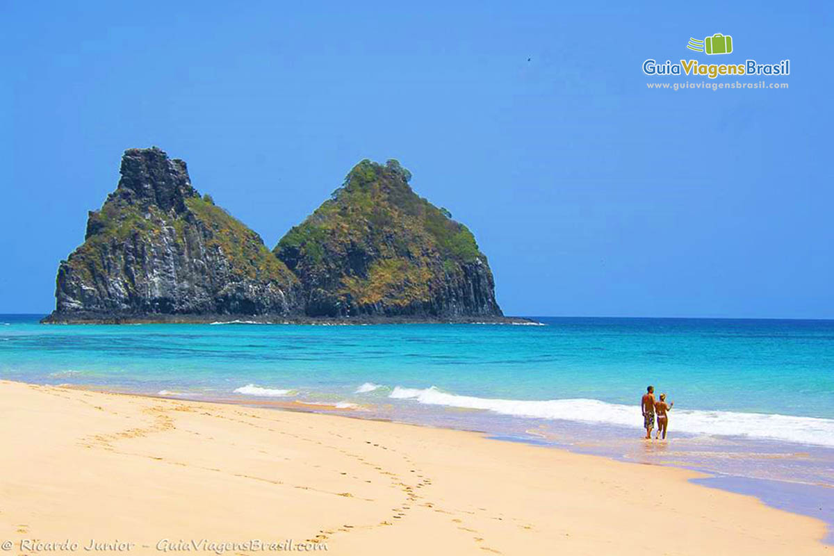 Imagem de dois turista parado na beira da praia olhando para horizonte e ao fundo o Morro Dois Irmãos, em Fernando de Noronha, Pernambuco, Brasil