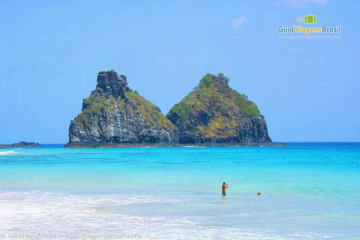 Imagem de dois turistas nas águas da praia e ao fundo o Morro Dois Irmãos, em Fernando de Noronha, Pernambuco, Brasil.