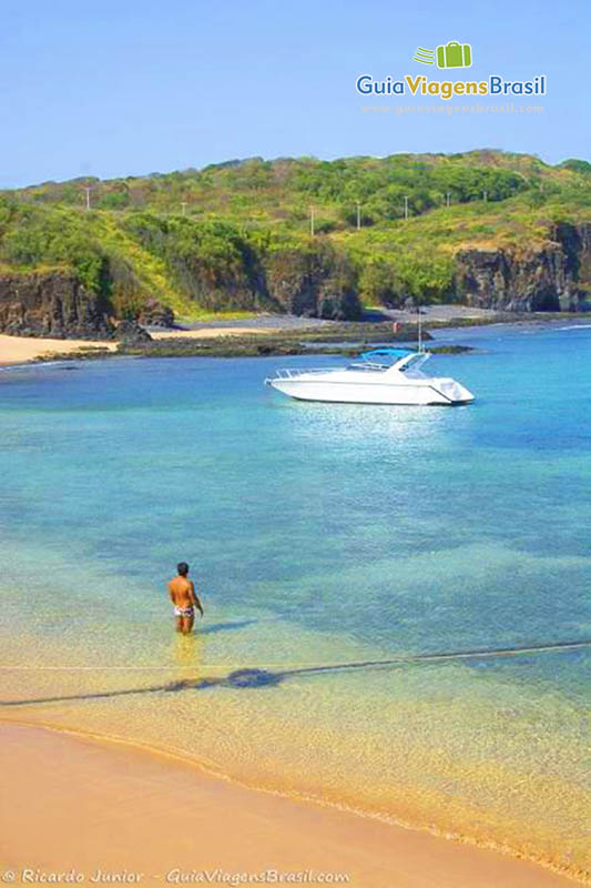 Imagem de turista na beira da praia olhando para uma linda lancha parada, em Fernando de Noronha, Pernambuco, Brasil.