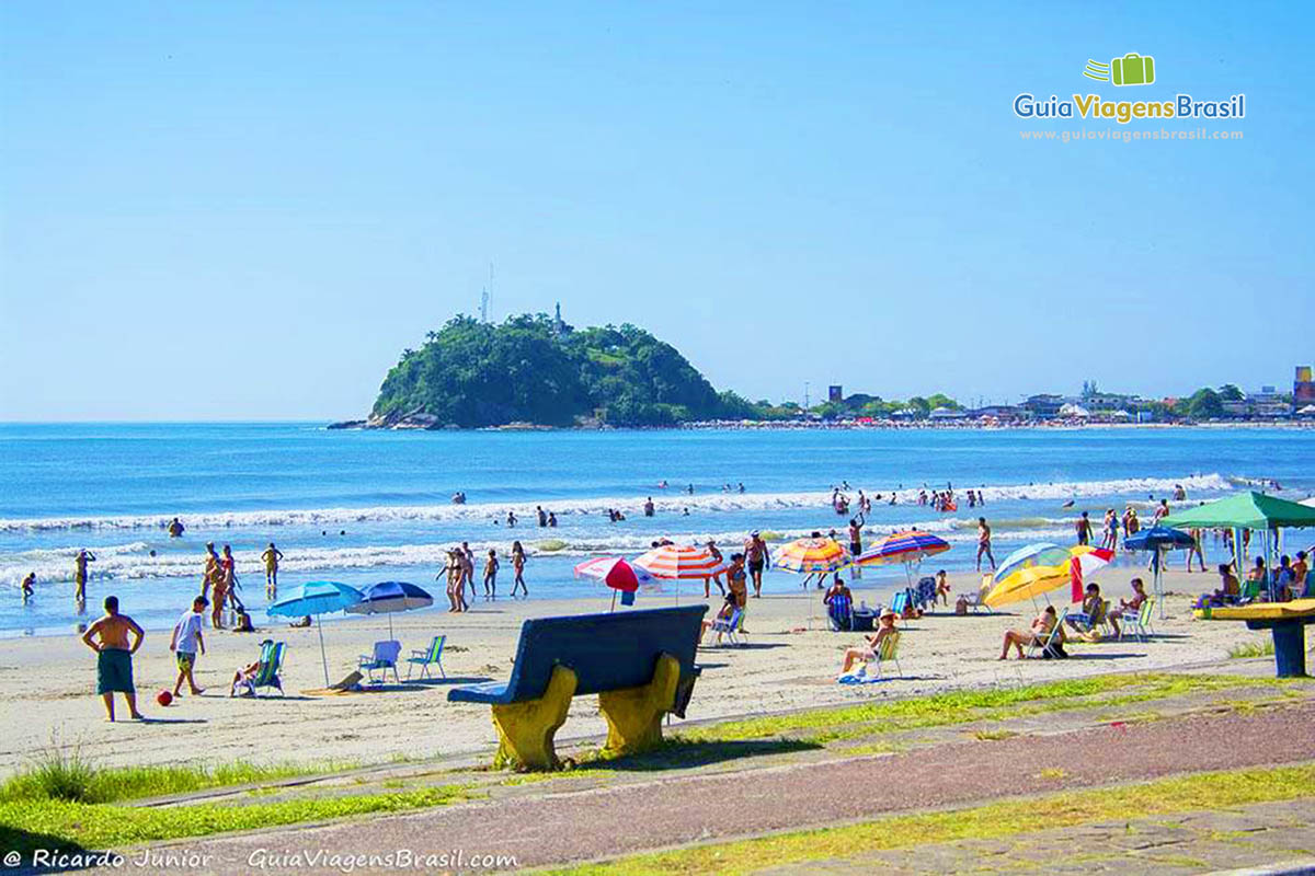 Imagem de banco de cimento na orla da praia e nas areias guarda-sol e ao fundo o mar da Praia de Guaratuba, em Santa Catarina, Brasil.