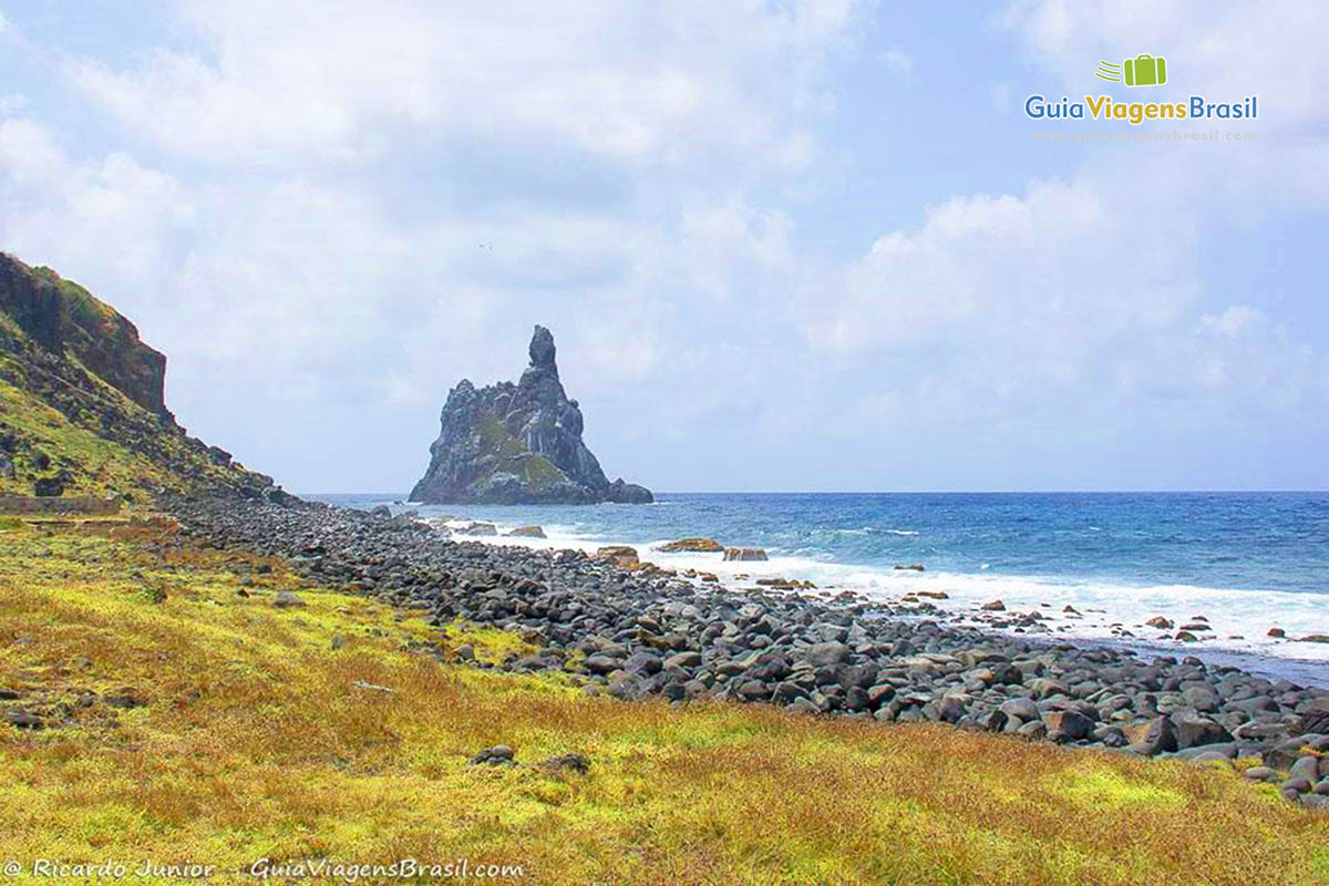 Imagem do canto da praia, com gramas, pedras e lindo mar com grande pedra no meio das águas, na Praia da Atalaia, em Fernando de Noronha, Pernambuco, Brasil. 