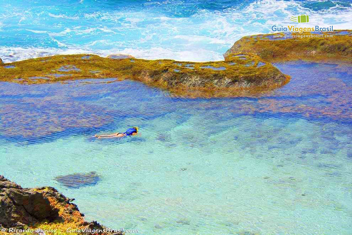 Imagem da piscina natural, as águas são transparentes que é possível ver os pé as pedras, tudo até a areia no fundo, na Praia da Atalaia, em Fernando de Noronha, Pernambuco, Brasil.
