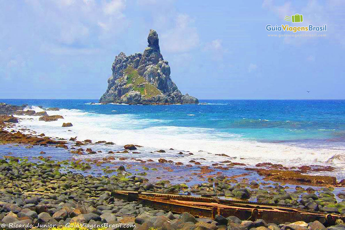 Imagem atrás da piscina natural, ondas batendo nas pedras e ao fundo uma pedra, na Praia da Atalaia, em Fernando de Noronha, Pernambuco, Brasil.