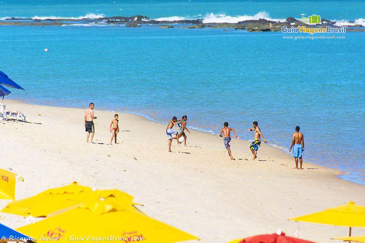 Imagem da Praia Barra de São Miguel, com adolescentes jogando bola na areia, aproveitando um dia ensolarado, em Alagoas, Brasil. 